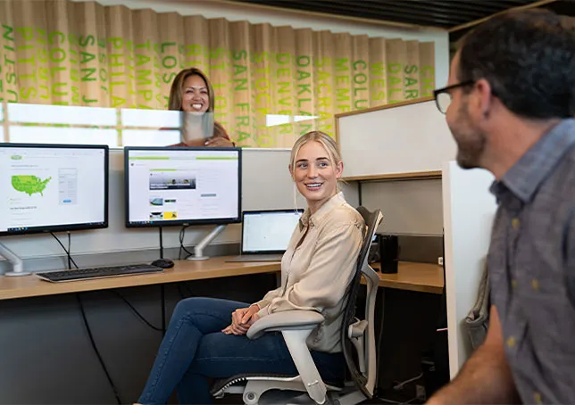 Woman smiling at computer desk with colleagues in office setting