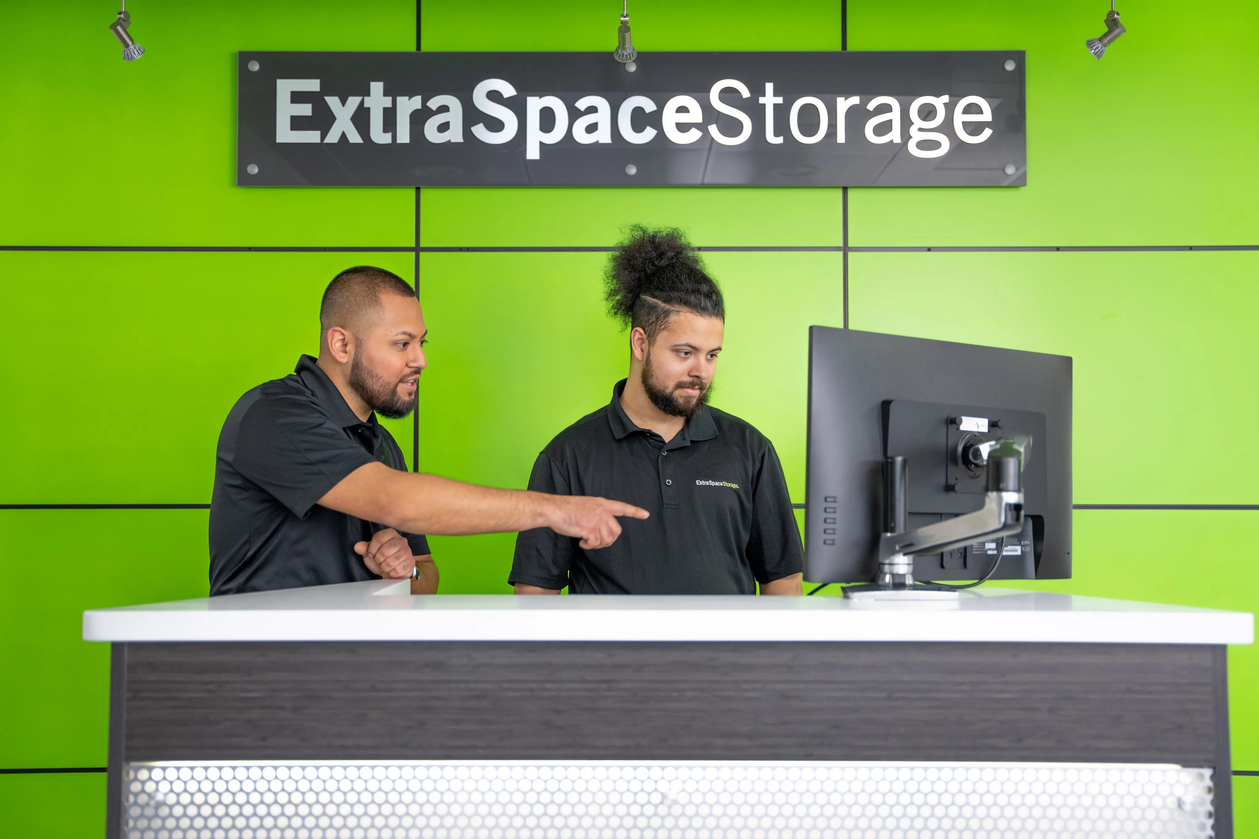One employee is pointing at a computer screen, while the other stands nearby. The background features a bright green wall with the ExtraSpace Storage logo prominently displayed