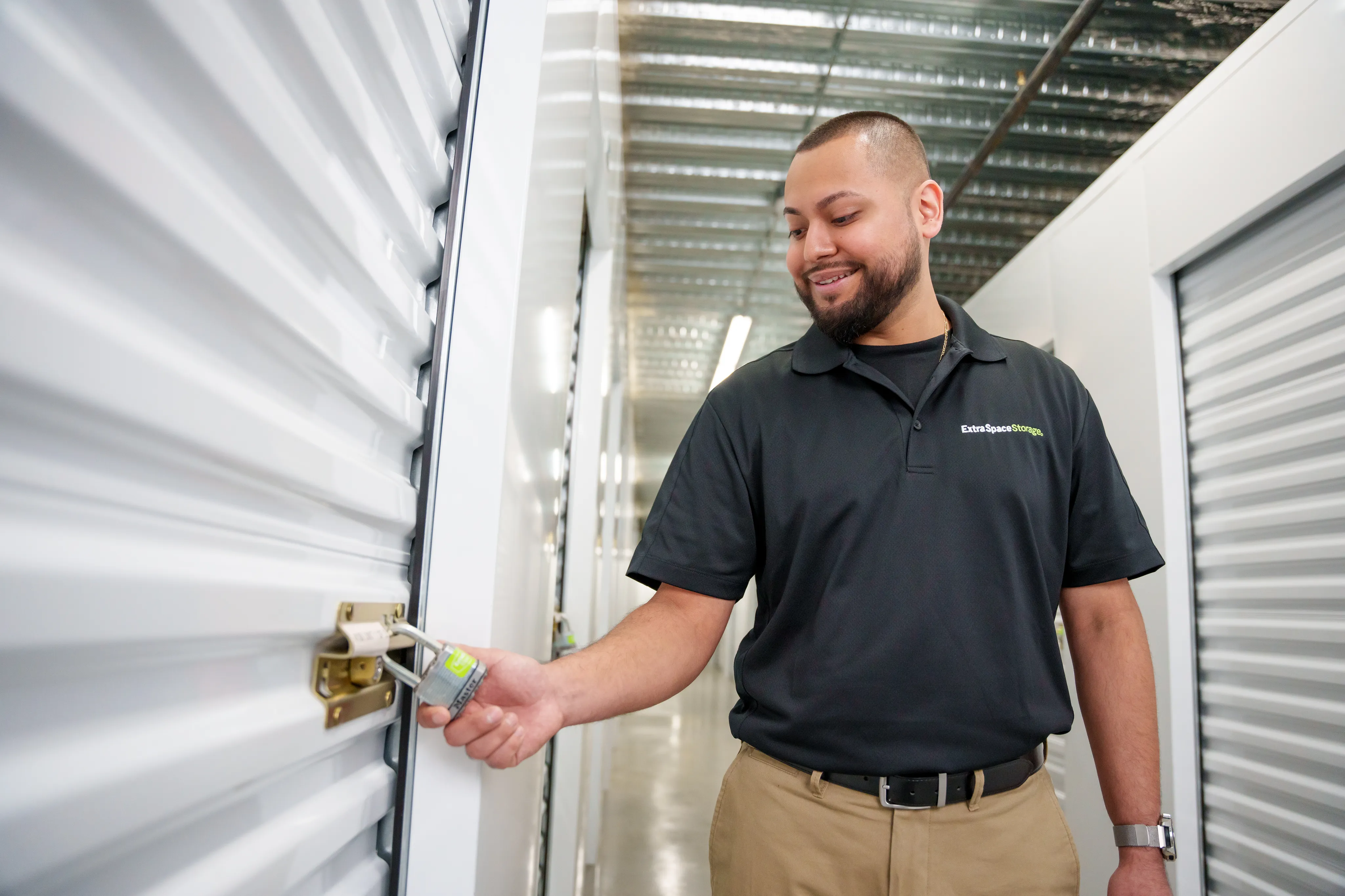 A person wearing a black polo shirt with the Extra Space Storage logo, unlocking a storage unit door with a padlock