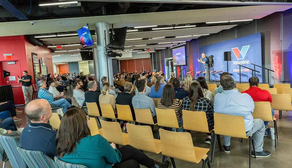 A wide shot of a speaker presenting on stage to a large group of seated employees at a corporate event.