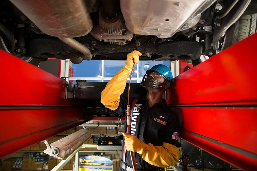 A **Valvoline technician** in a helmet and safety mask is servicing a car from the pit underneath.
