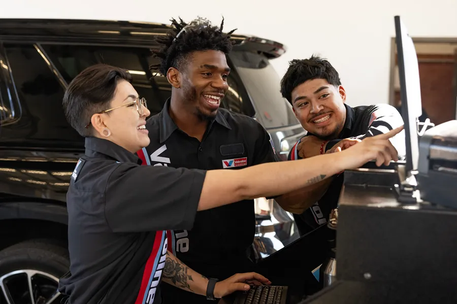 Three cheerful Valvoline technicians are collaborating and smiling while working on a laptop next to a vehicle.