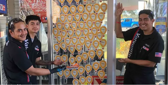 Three smiling Valvoline service center team members are standing with a donation wall covered in heart-shaped notes.