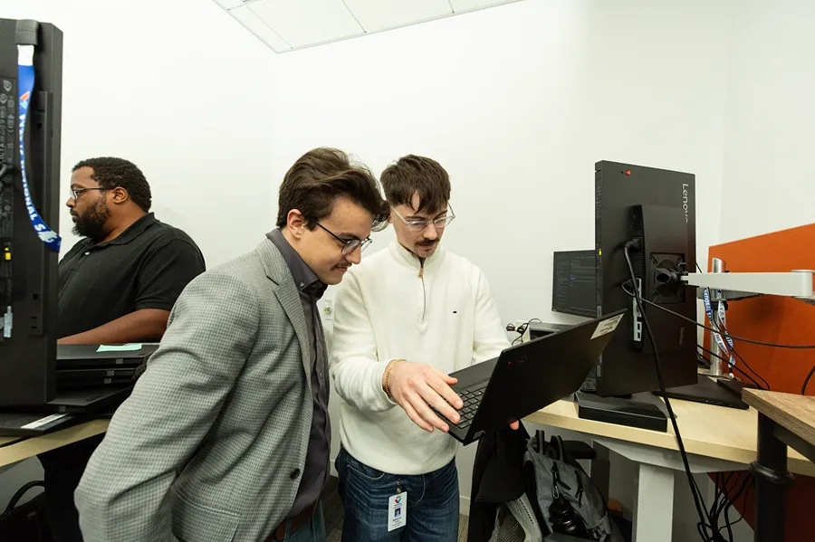 Two male colleagues are collaborating and looking at a laptop screen in a bright office environment.