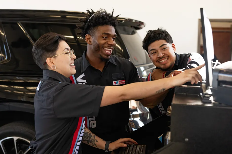 Three cheerful **Valvoline technicians** are collaborating and smiling while working on a laptop next to a vehicle.