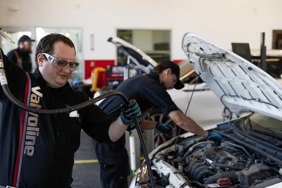 A focused **Valvoline technician** in safety glasses is working under the hood of a car.