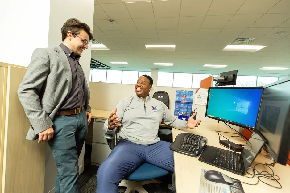 Two laughing male colleagues are interacting at an office desk with multiple computer screens.