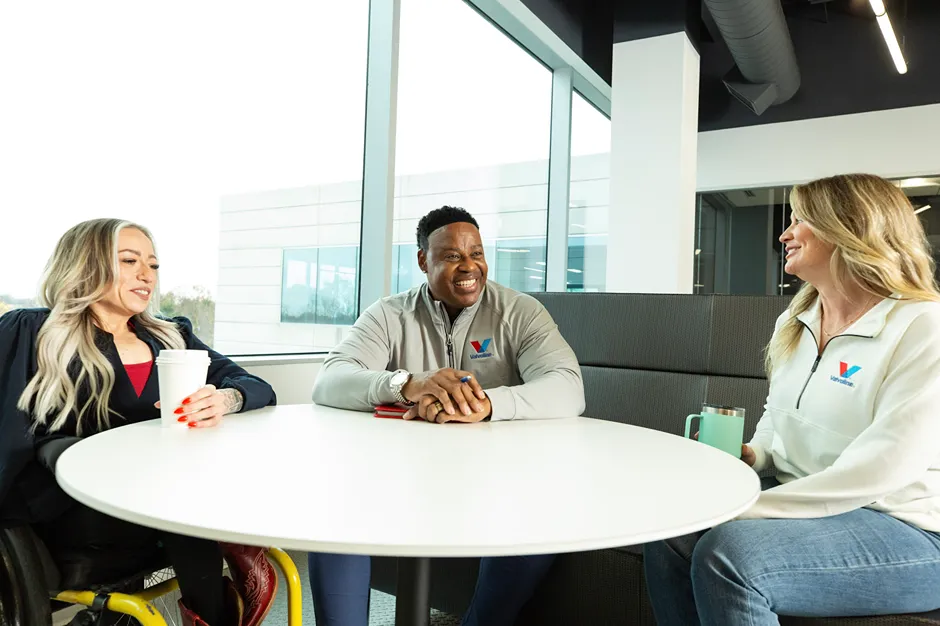 Three diverse colleagues are smiling while having a casual discussion at a white table.
