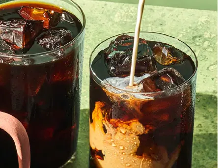 Two glasses of iced coffee with milk being poured into one glass on green surface
