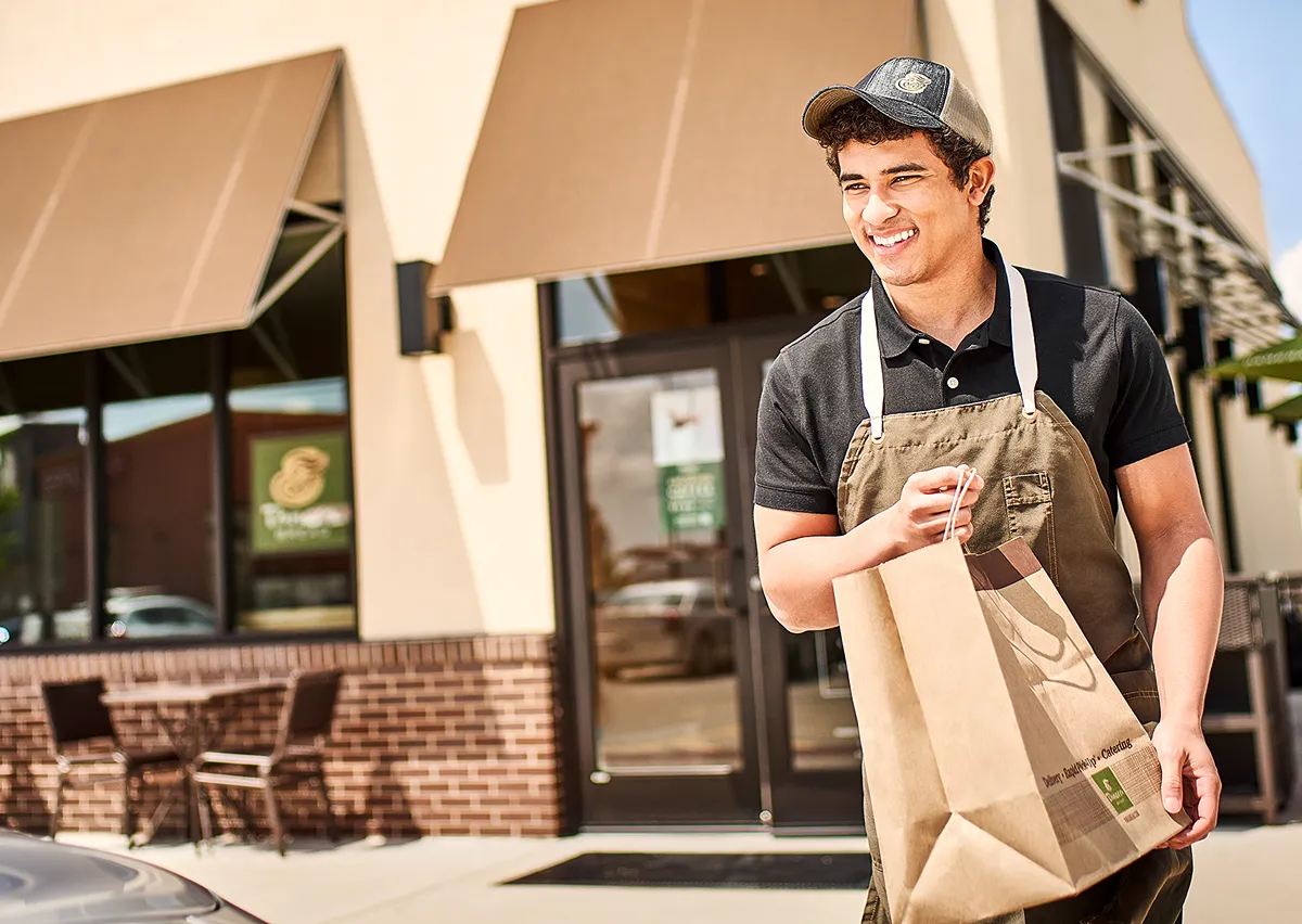 Smiling worker in apron carrying brown bag outside store