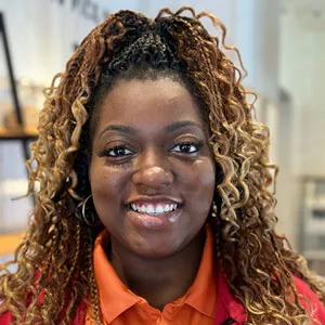 A smiling person with curly hair wearing an orange shirt indoors.
