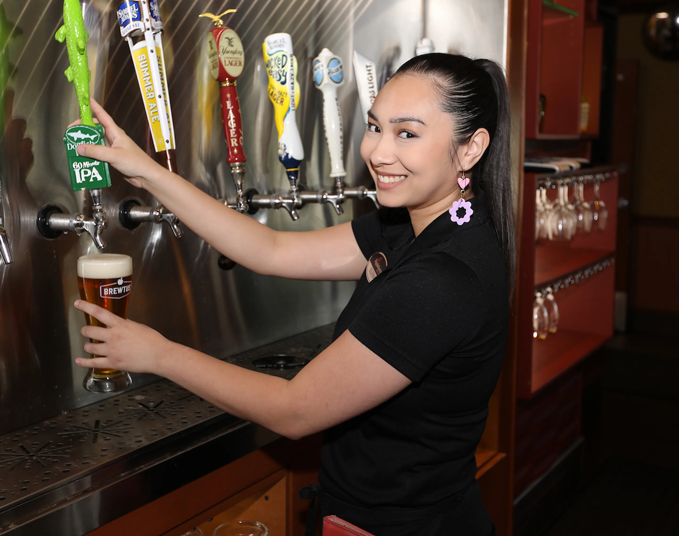 A bartender pouring a beer from a tap at a bar, holding a glass of Brewtits IPA under a row of colorful beer taps