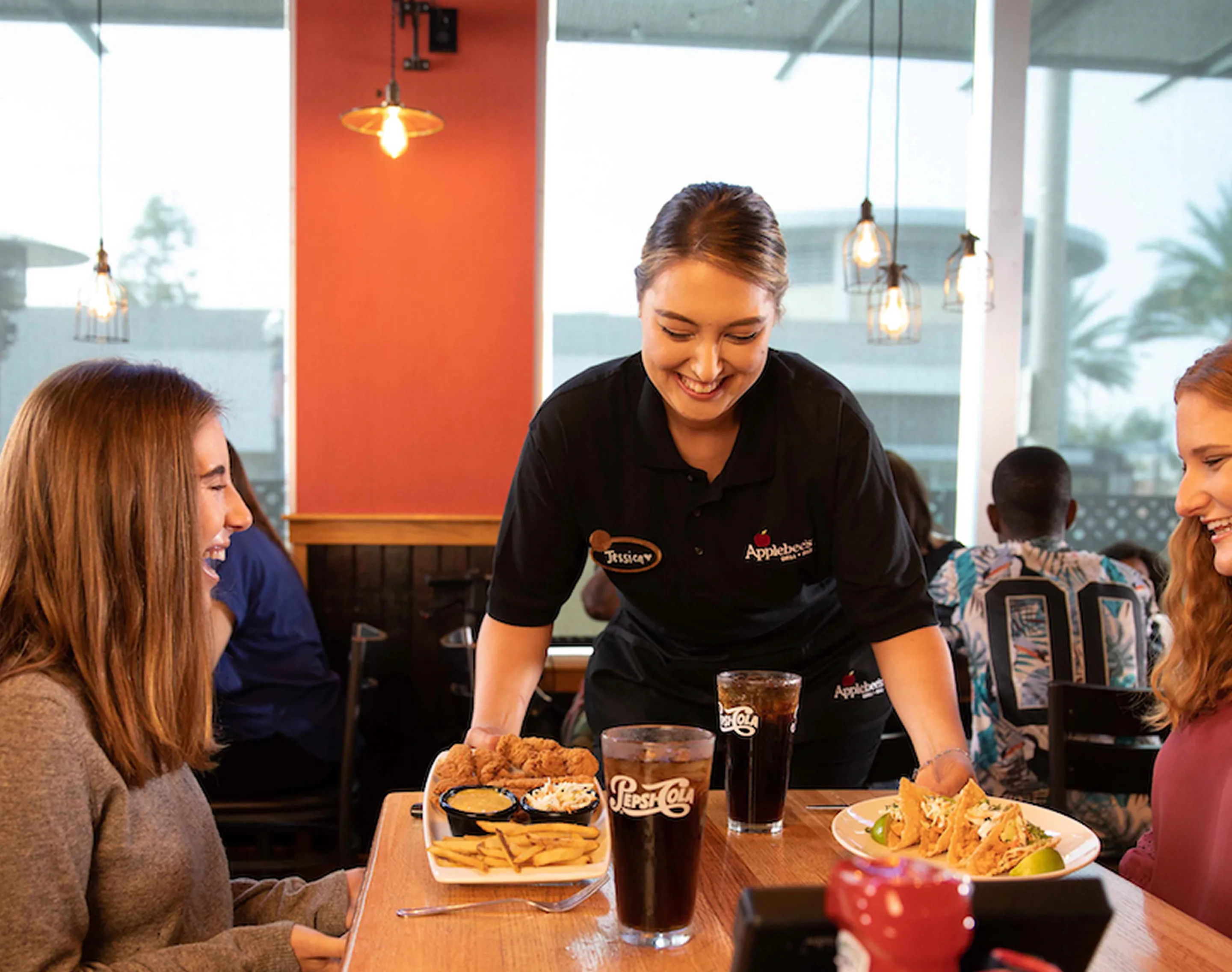 A server in a black uniform with an Applebee's logo is smiling while serving customers in a brightly lit restaurant with orange walls, hanging pendant lights, and large windows showing a view of palm trees outside
