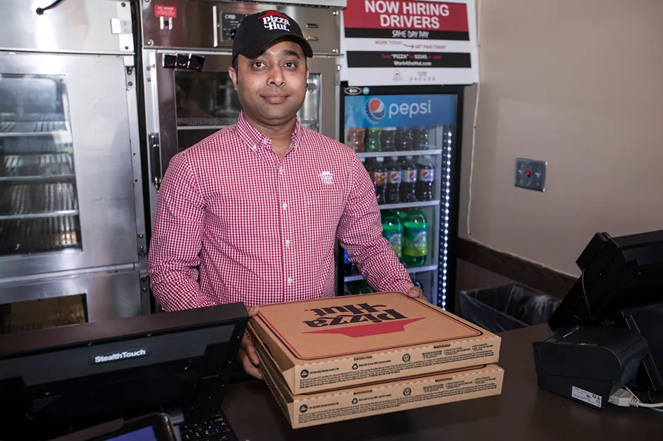 A Pizza Hut employee in a red and white checkered shirt and a black logo cap stands behind a counter, smiling at the camera.