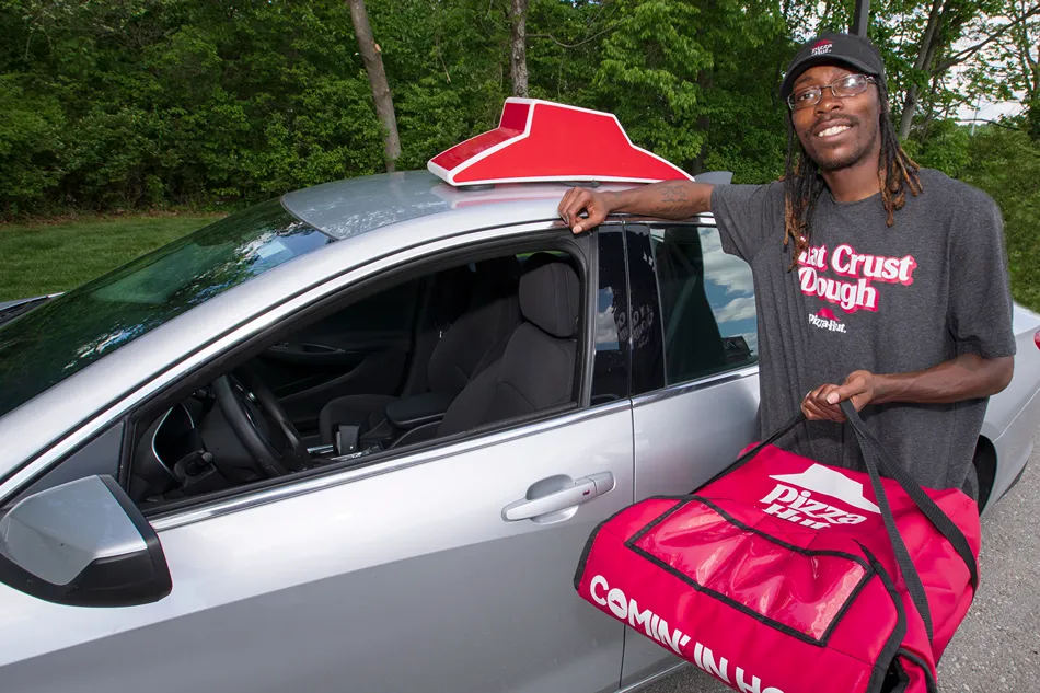 A man in a gray shirt with the words Eat Crust Dough on it is standing next to a silver car, holding a red pizza delivery bag.