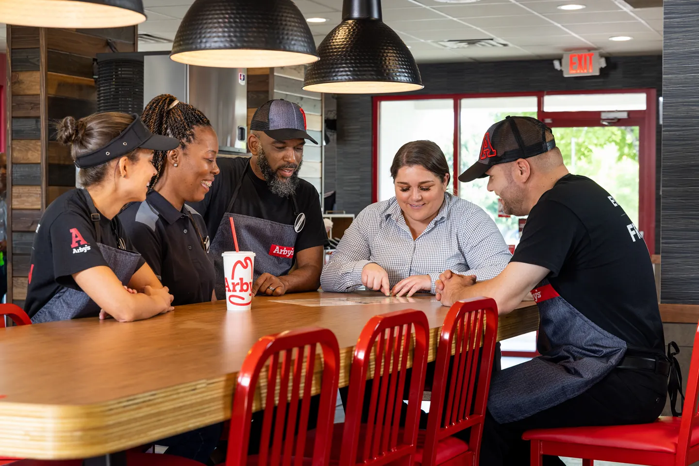 Five diverse Arby's employees, some in uniform, smiling and engaged in a discussion around a wooden table with red chairs in a restaurant setting.