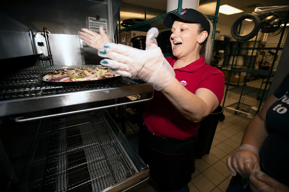 A woman in a red uniform is taking a pizza out of an oven wearing protective gloves.