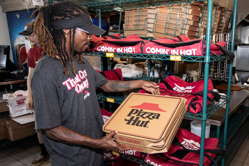 Pizza Hut team member wearing 'That Crew Energy' shirt selecting pizza boxes from a rack filled with insulated delivery bags in a busy kitchen environmen