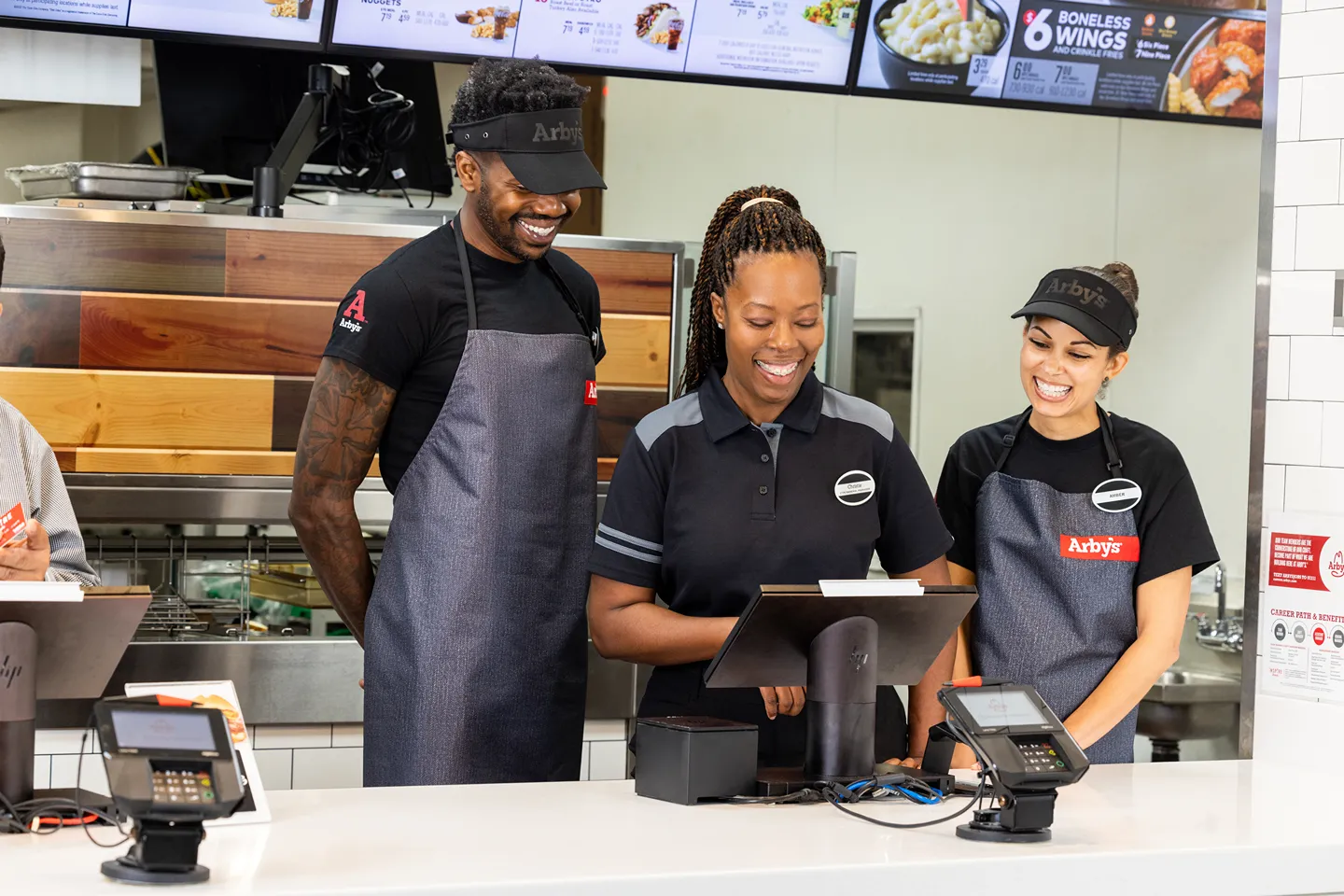 Three happy Arby's team members, two women and one man, wearing uniforms and aprons, smiling while working behind the counter with a POS system.