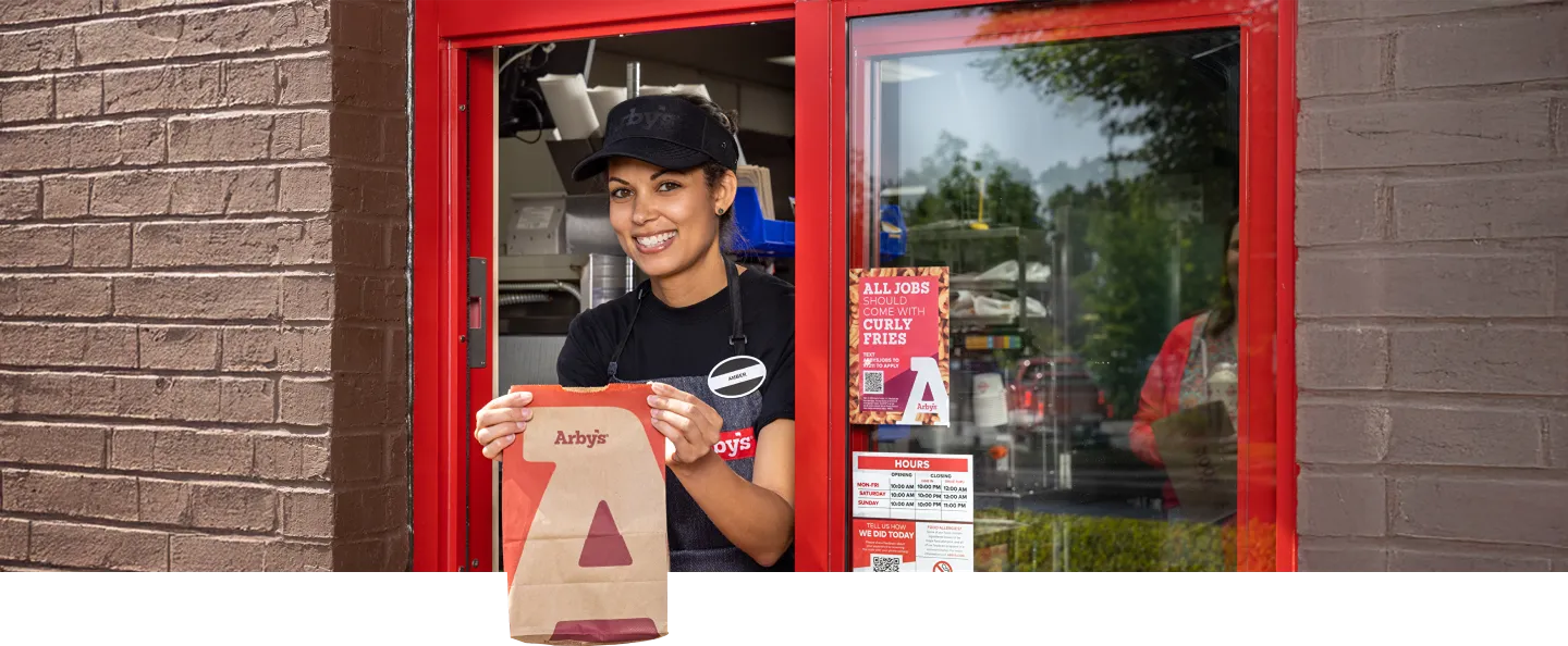 A smiling female fast-food worker in a black cap and apron handing an Arby's take-out bag through a drive-thru window