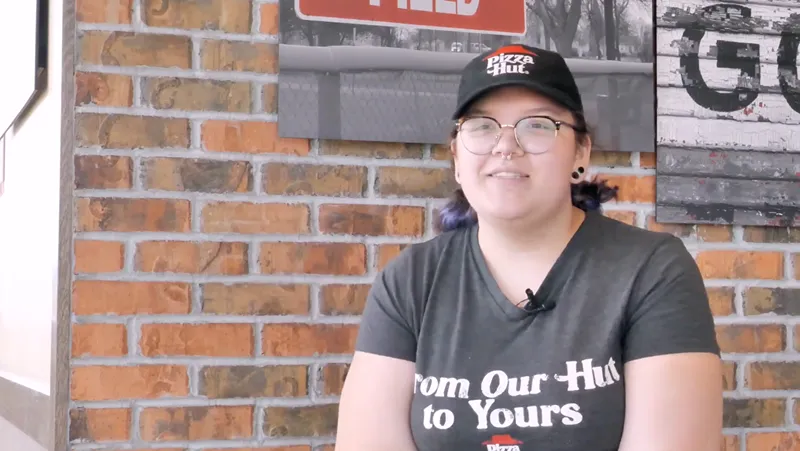 A young woman wearing a Pizza Hut uniform and glasses is standing in front of a brick wall.