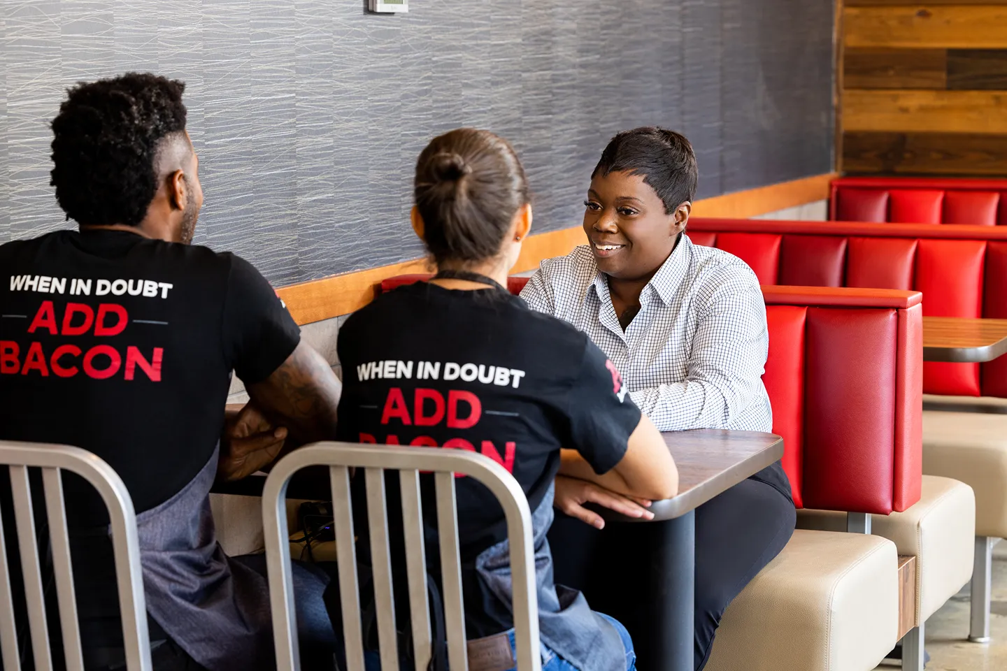 Three people in a restaurant booth, two wearing 'Add Bacon' shirts, having a discussion.