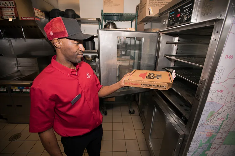 A person in a red uniform is taking a pizza box out of a commercial oven.