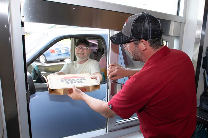 A man in a car is receiving a pizza order from a Pizza Hut employee at a drive-thru window.