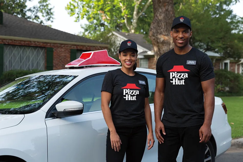 Two Pizza Hut employees, a woman and a man, stand next to a white car.