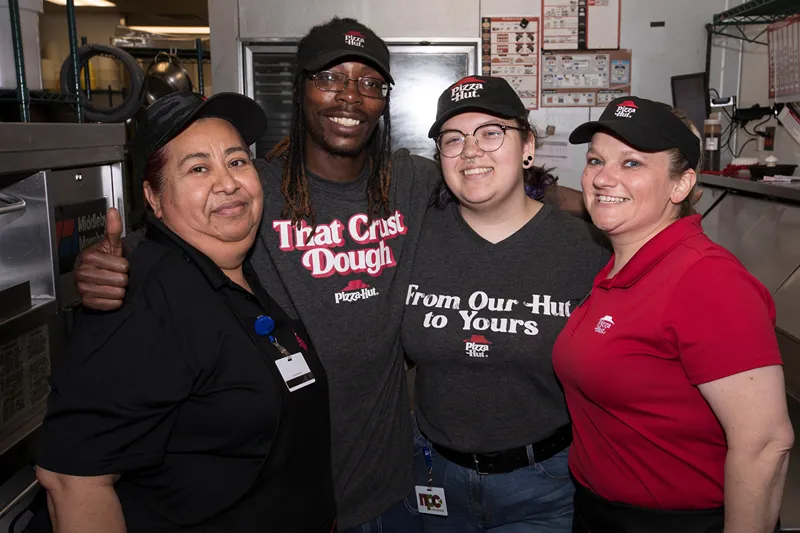 A group of four people, likely employees of a Pizza Hut, are posing for a photo, smiling and appearing friendly.