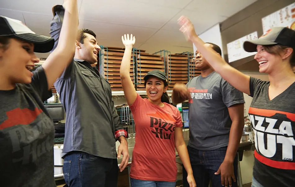 Pizza Hut team members smiling and giving high fives in a kitchen, showing teamwork and enthusiasm.
