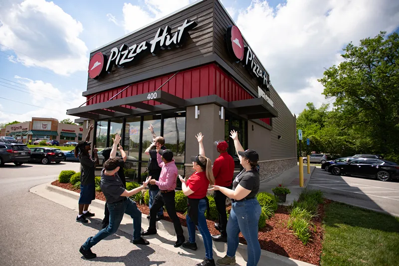 Group of enthusiastic Pizza Hut team members outside the restaurant, smiling and raising hands toward the building under a sunny sky.