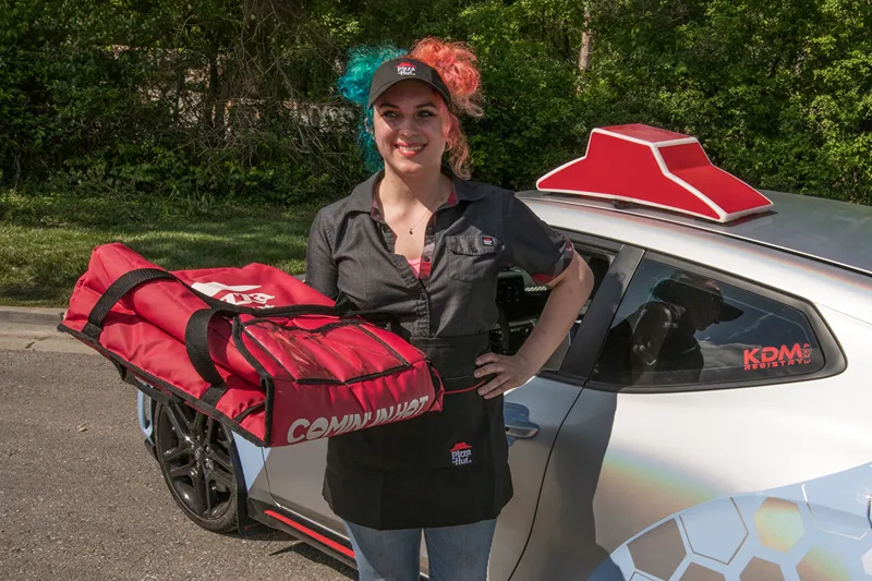 Smiling Pizza Hut delivery driver with red and blue hair holding insulated pizza bag next to branded delivery car in a residential neighborhood.
