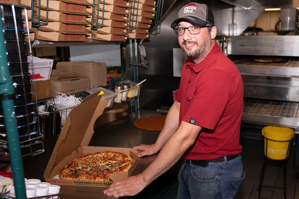 A man in a red Pizza Hut uniform is preparing to hand over a pizza in a cardboard box.
