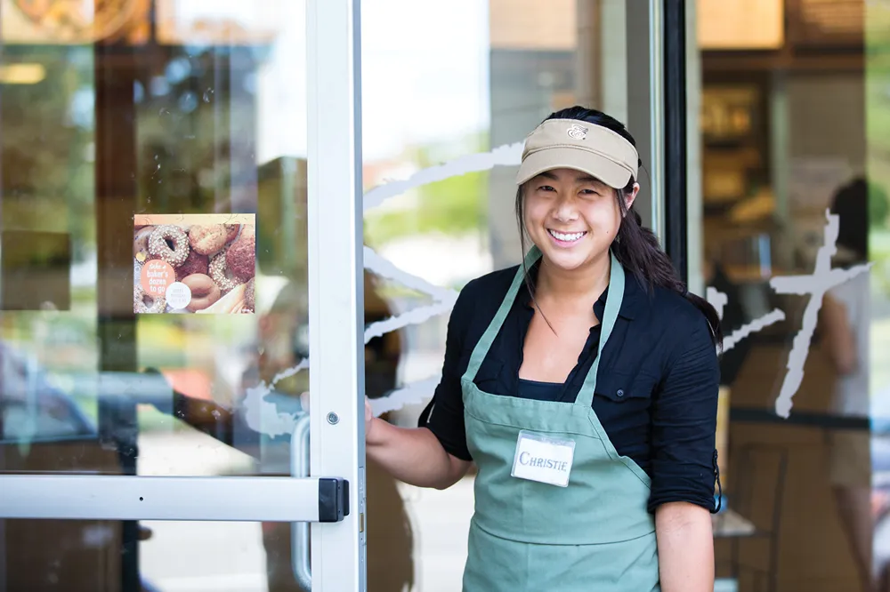 Smiling woman standing at a bakery door wearing an apron and name tag that says Christie