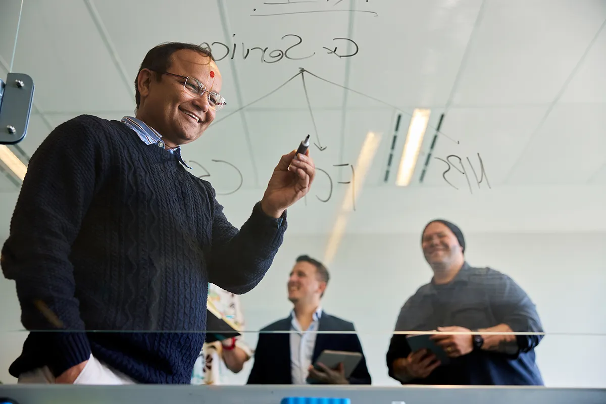 A man writes on a transparent surface, two men standing behind him in the background
