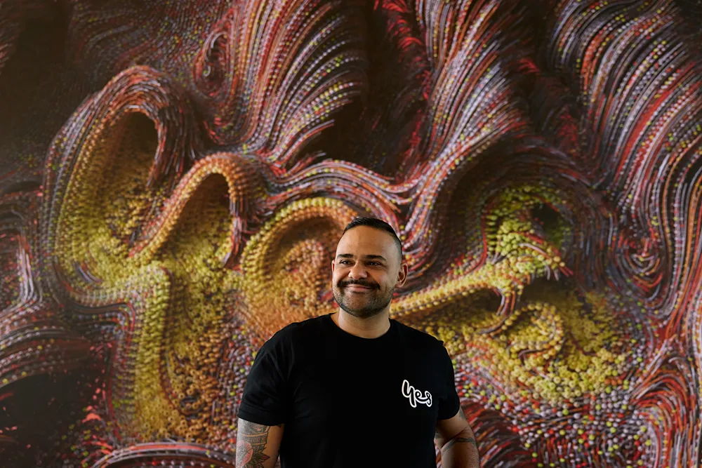 A smiling man with a beard and tattoos wearing a black 'Yes' t-shirt, standing in front of a vibrant, abstract, swirling artwork