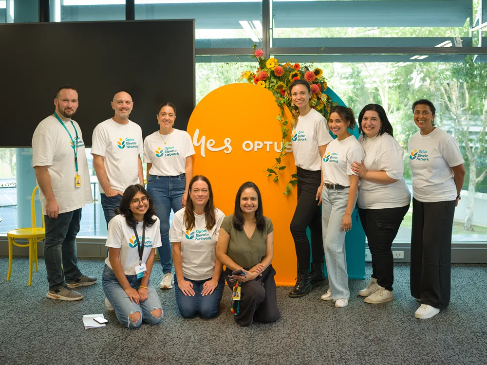 A group of diverse men and women in white Optus t-shirts smiling around a 'Yes Optus' sign