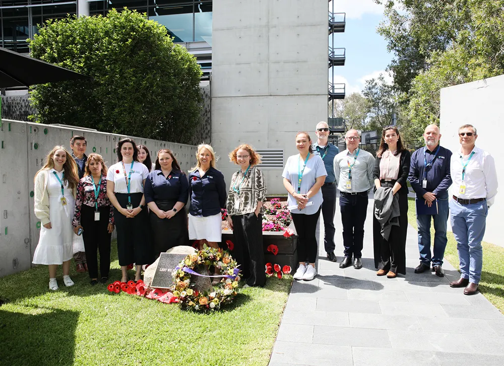 A group of diverse individuals standing respectfully around a memorial wreath in a corporate outdoor setting on a sunny day