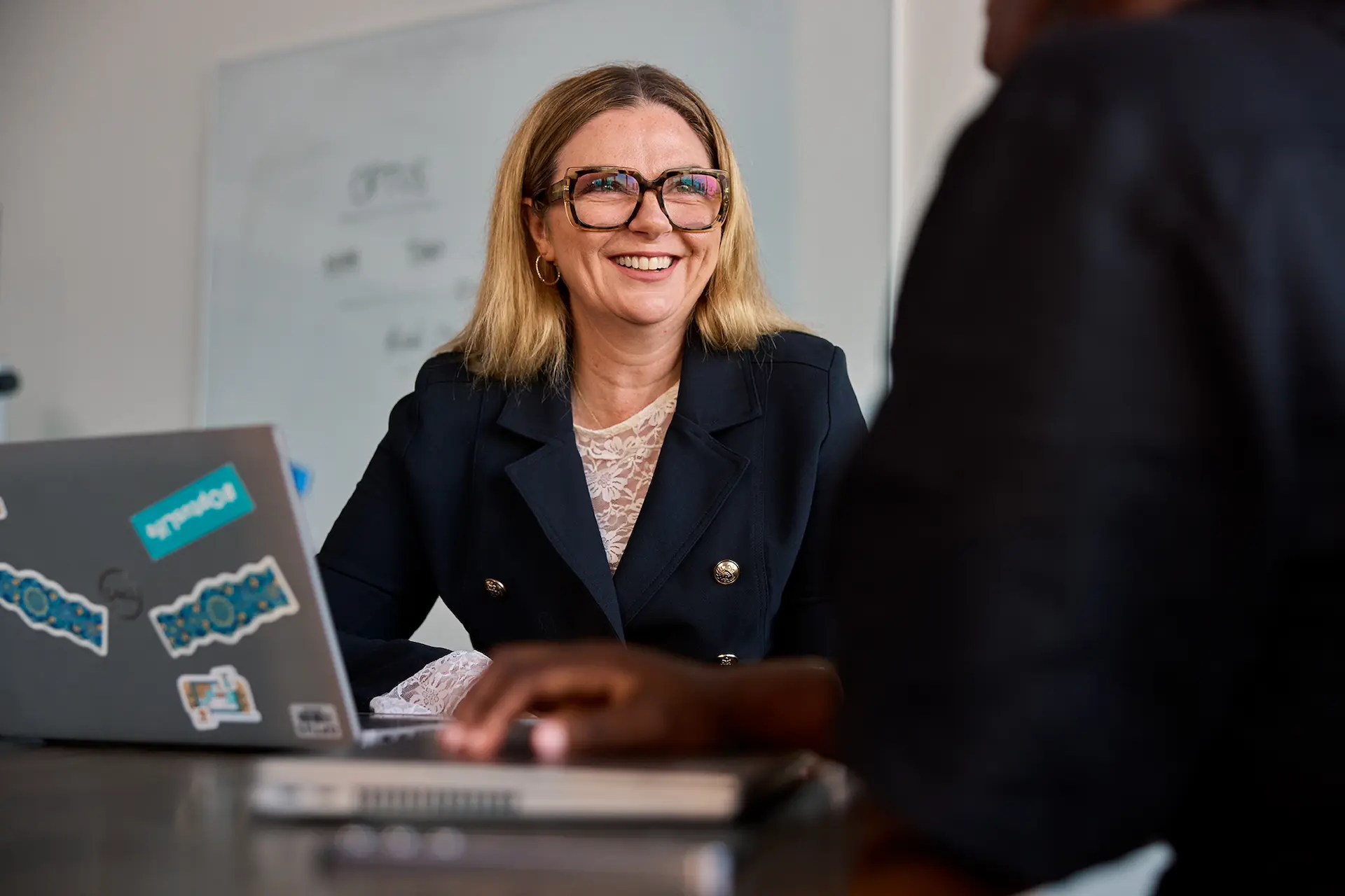 A woman wearing glasses and blue suit jacket sitting at a table smiling