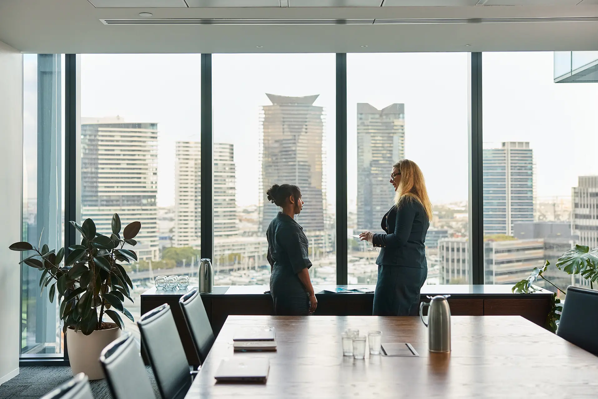 Two women standing in a office conference room with an urban skyline behind them