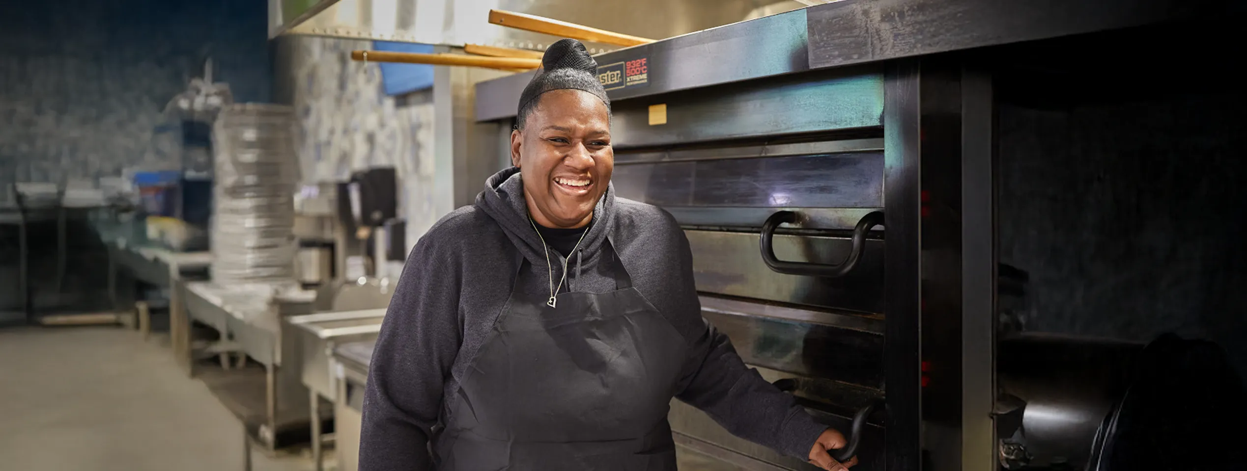 Woman wearing apron in a commercial kitchen smiling by large industrial oven.