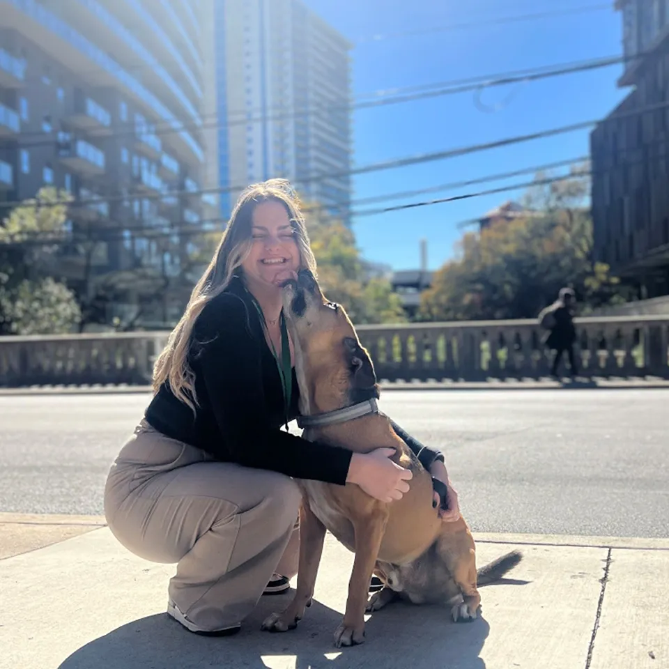 Camryn, HRIS Business System Analyst, smiles while being kissed by her dog outdoors, with city buildings in the background.