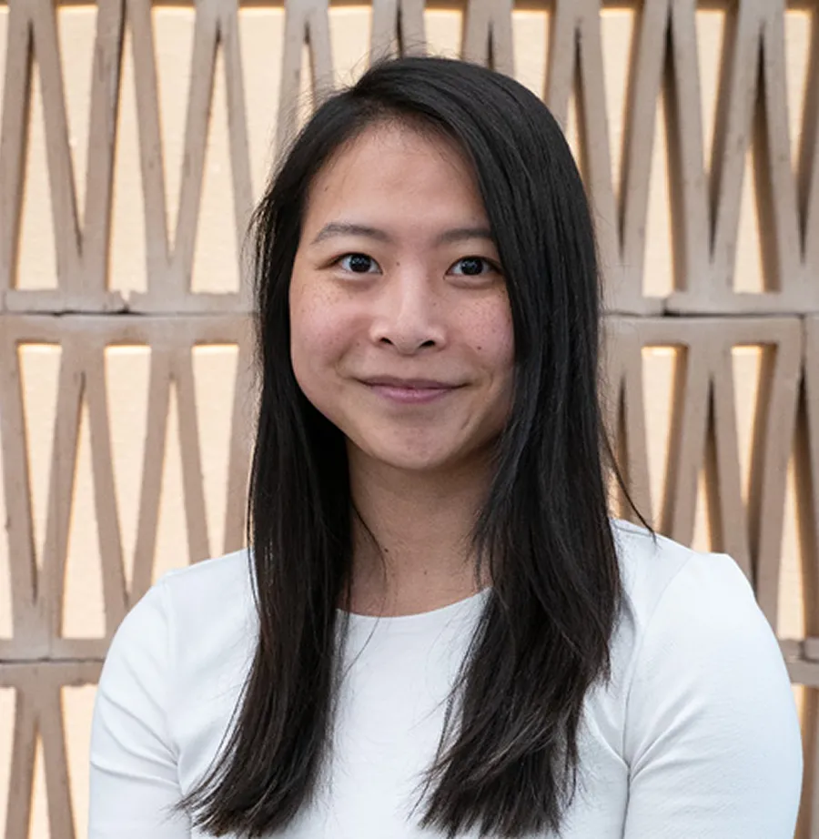 Smiling Asian woman with long black hair wearing a white shirt in front of a geometric wall.