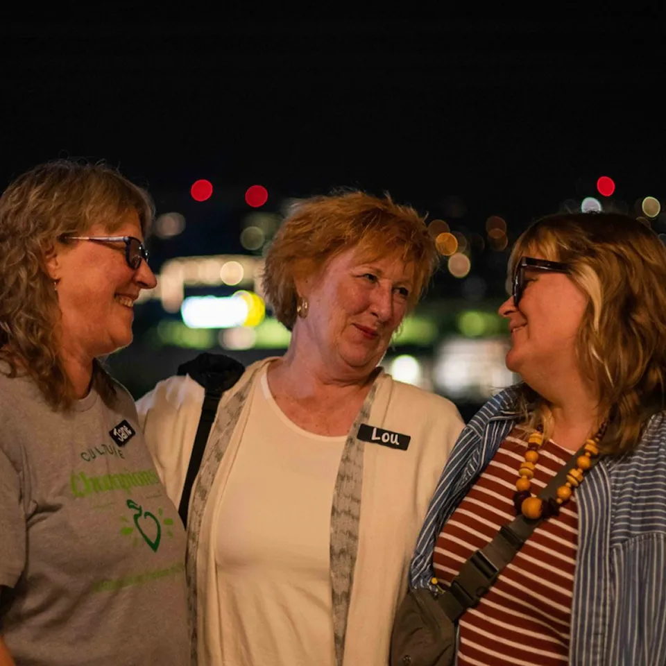 Three women smiling and looking at each other with blurred city lights in the background, one woman labeled Lou.