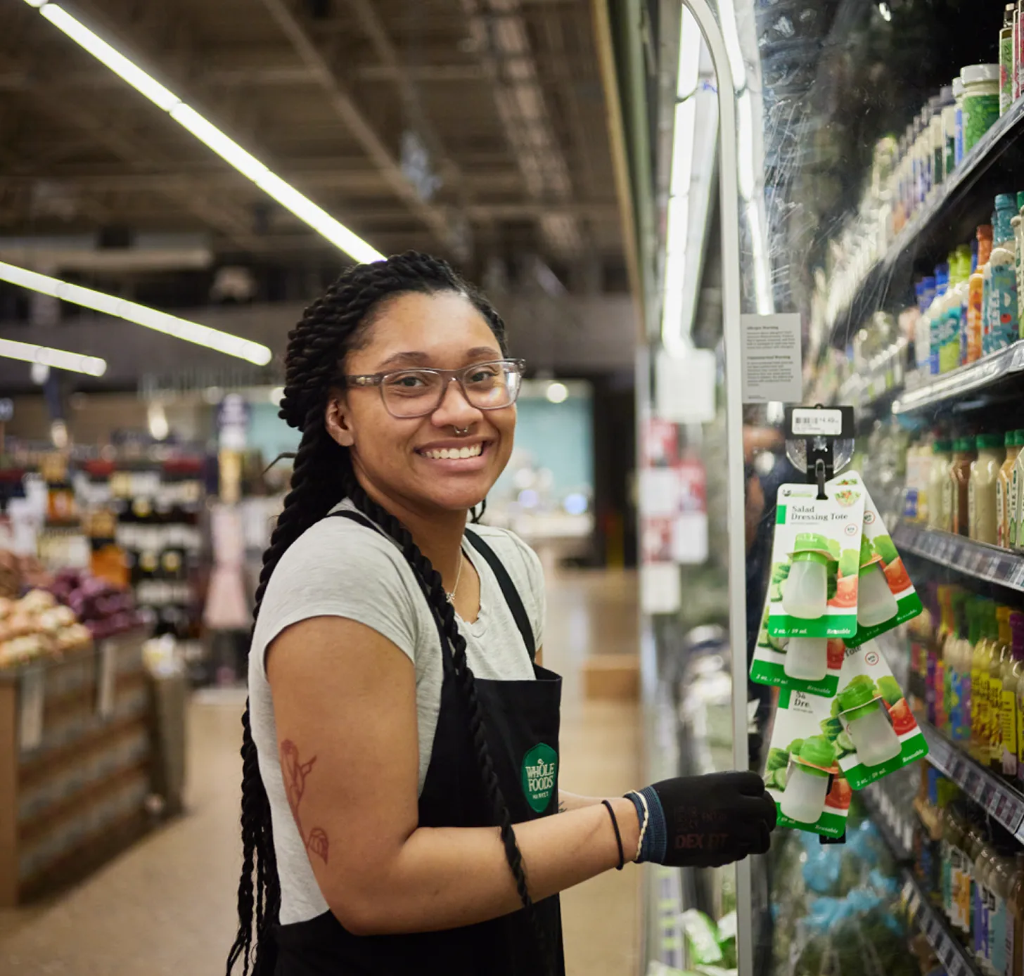 Smiling Whole Foods Market employee wearing a black apron and gloves standing in front of a refrigerated display case.