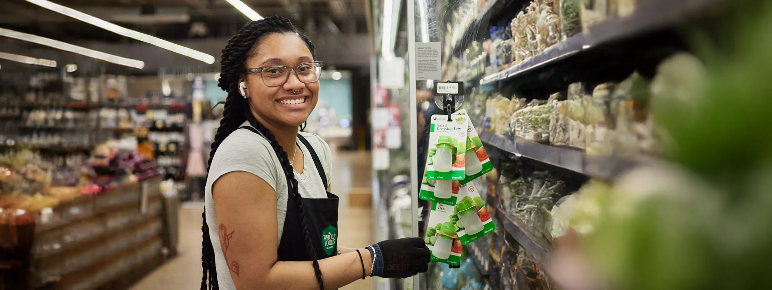 A smiling Whole Foods Market team member with dreadlocks and glasses wearing an apron and black gloves restocking salad dressing on a shelf in a store aisle.