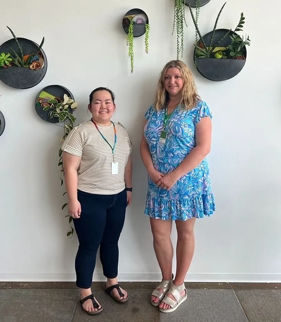 Two women smiling in front of a white wall with decorative planters, one woman is likely Courtney, Manager.