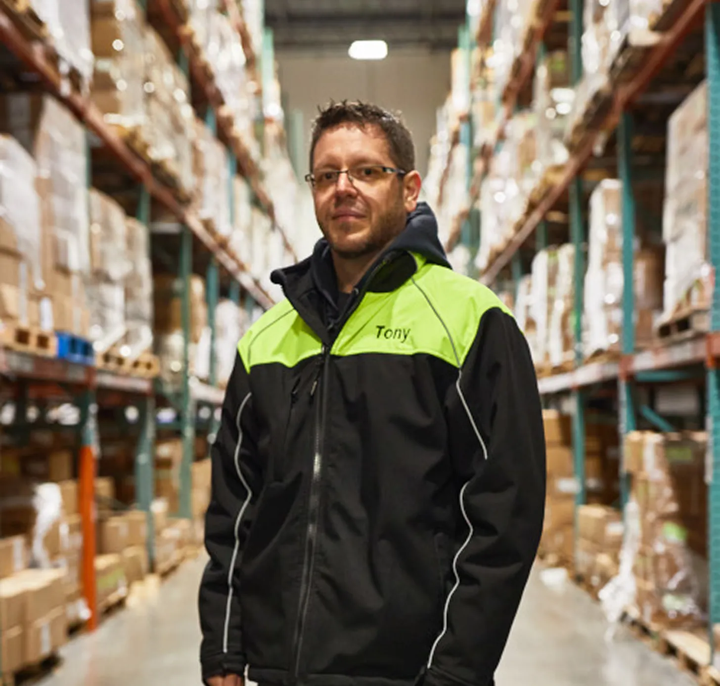 Warehouse worker in a black jacket with neon green shoulders, standing in a warehouse aisle with stacked boxes on shelves.