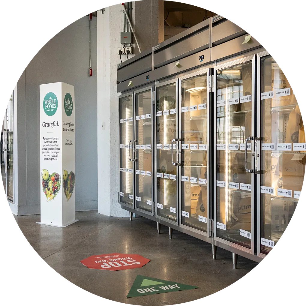 Whole Foods Market refrigeration units with customer order pickup bags visible, including floor signage for one way and stop, plus a branded display.